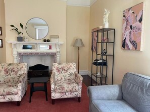Un salon avec une cheminée, deux fauteuils à motifs floraux, un canapé gris, une étagÚre en métal noir et un tableau accroché au mur.
