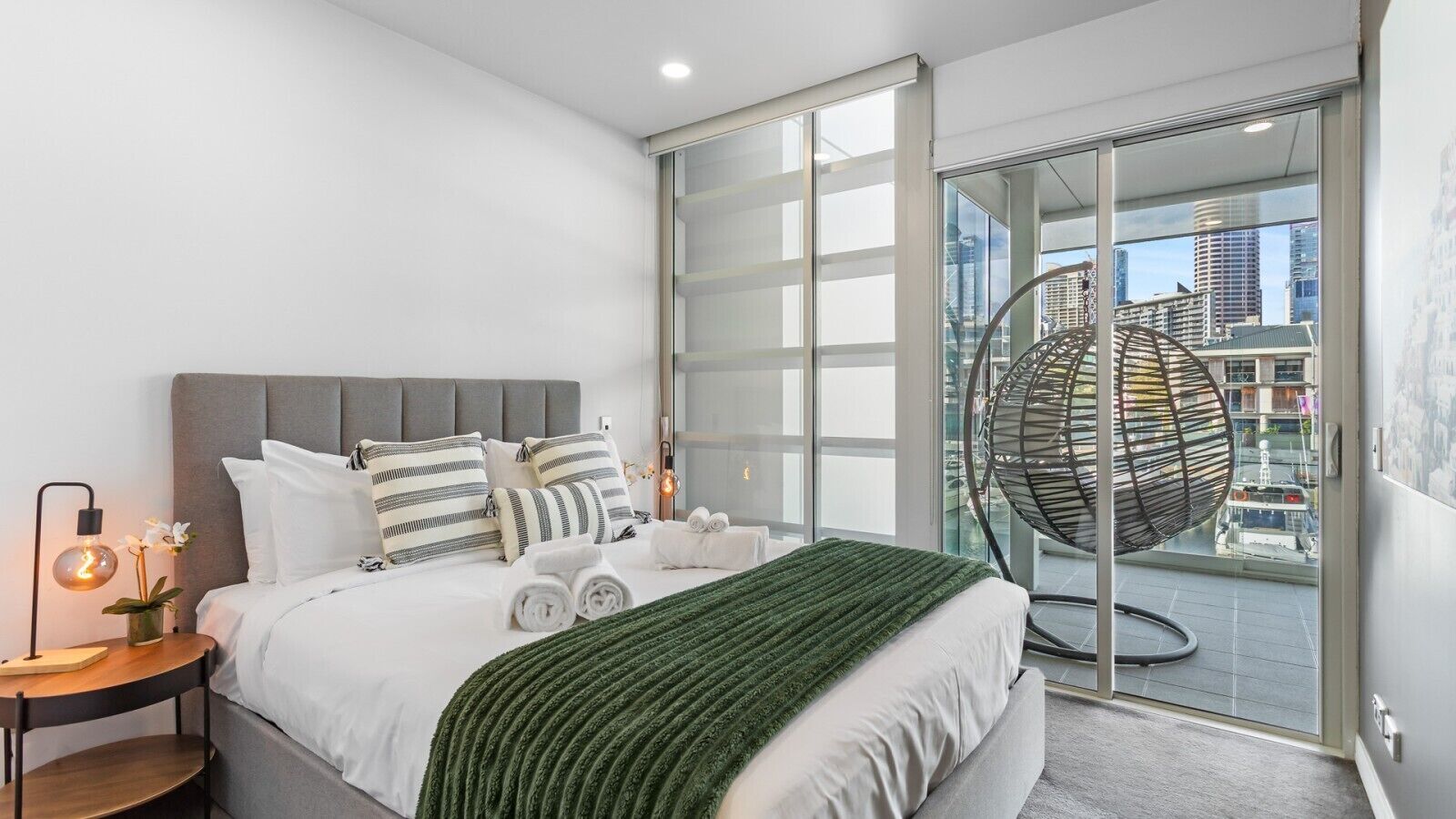 Ein modernes Schlafzimmer mit einem Bett, Kissen, einem Nachttisch mit Lampe und einem großen Fenster mit Blick auf die Stadt.