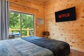 Een houten kamer met een bed, een aan de muur bevestigde televisie, een raam met uitzicht op bomen en een stoel.