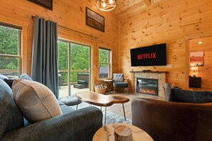 A cozy cabin interior with a fireplace, a flat-screen TV, and a large window overlooking greenery. - Blue Sky Retreat.  Cozy 2 bedroom cabin in Red River Gorge. (Beattyville)