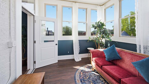 Ein sonnendurchfluteter Raum mit einem roten Sofa, einem kleinen runden Tisch und einem großen Fenster mit Vorhängen.