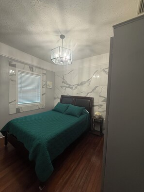 Un dormitorio con cama, mesita de noche, lámpara de pared y ventana con persianas.