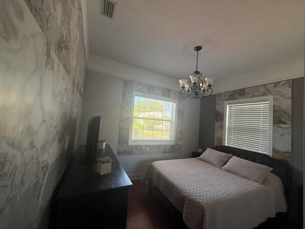 A bedroom with a bed, a dresser, a window with blinds, a chandelier, and a wall with a marble-like texture.