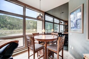 A dining area with a round table and chairs, large windows with a view of a forest, and a stone wall. - Beautiful Keystone Condo: Perfect Leaf Peeping! (Keystone)