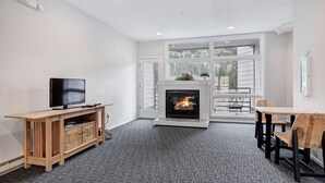 A modern living room with a fireplace, a television, a dining area, and a large window. - Beautiful Keystone Condo: Perfect Leaf Peeping! (Keystone)