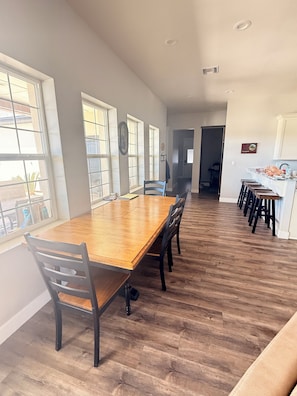 Un vaste espace de salle à manger avec une longue table en bois et des chaises, de grandes fenêtres et un coin cuisine doté de meubles blancs.