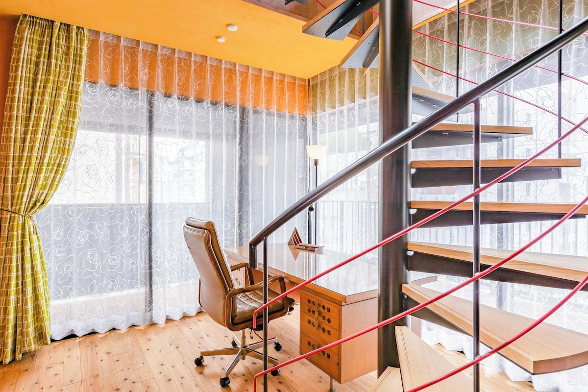 Un intérieur moderne avec un bureau en bois, une chaise, un escalier doté d’une rampe en métal et des rideaux légers.