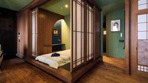 Um quarto em estilo tradicional japonês, com piso de tatame, cama baixa em plataforma, portas deslizantes e uma pequena prateleira com espelho e moldura de quadro.