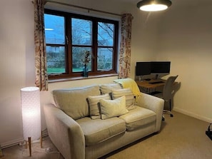 Ein Wohnzimmer mit einer Couch, einem Schreibtisch mit Computer, einer Lampe und einem Fenster mit Vorhängen.