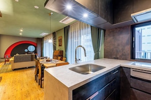 A modern kitchen with a large island, a dining area with a sofa and chairs, and a view of the city through large windows. - Spacious & Serene 84 ㎡ Retreat near Dotonbori, Kuromon Market & Shinsaibashi (大阪市)