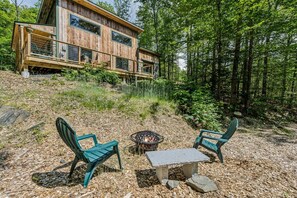 A wooden cabin with a deck, two chairs, a table, and a fire pit. - Gratitude Cottage, Quiet Nature Escape (Strafford)