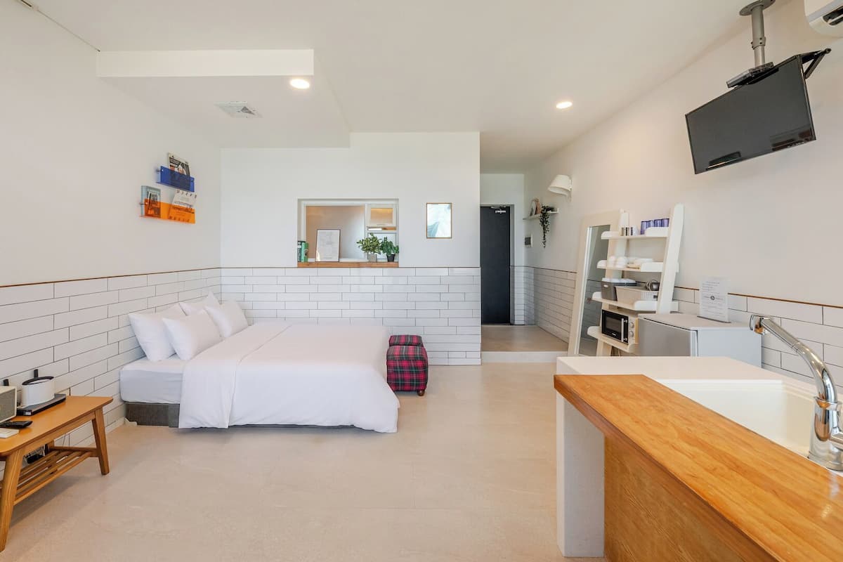 Habitación de hotel con cama, mesita de noche, un taburete pequeño, un banco de madera, televisor montado en la pared, zona de cocina con microondas y un lavabo con grifo moderno.