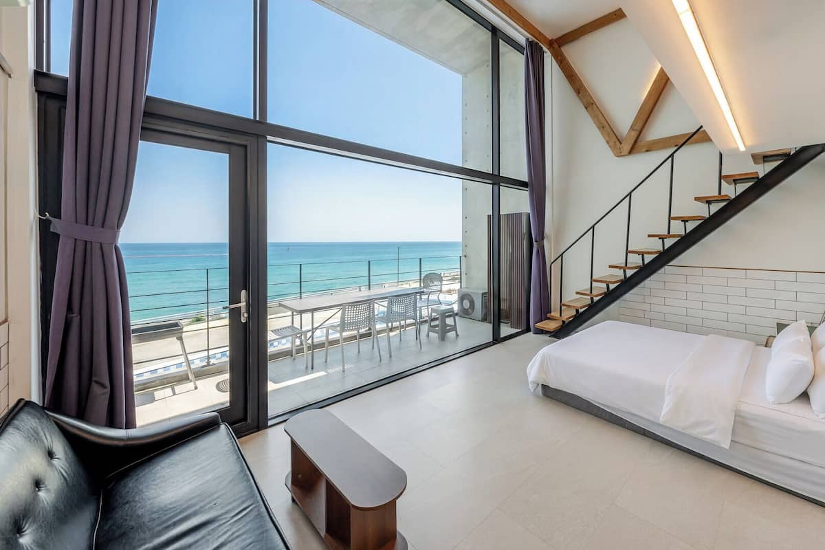 Un dormitorio moderno con un amplio ventanal con vistas al mar, una cama, una escalera y una zona de estar.