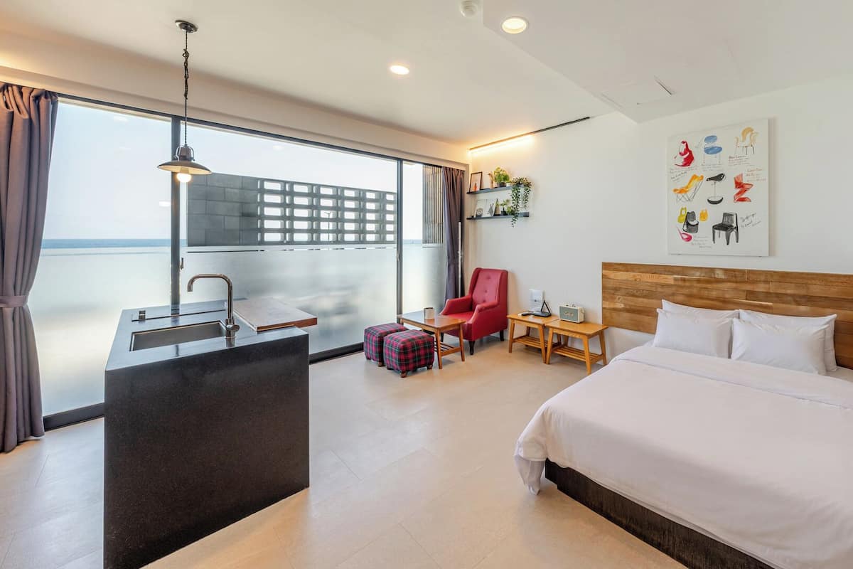 Habitación de hotel con cama, escritorio con lavamanos, una silla roja, una mesita y un ventanal con vistas.