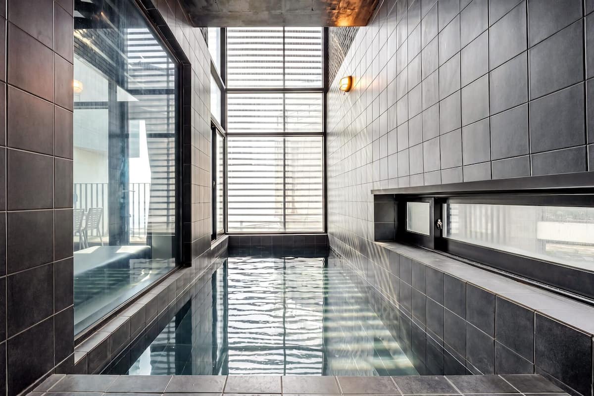 Piscina interior moderna con paredes de azulejos oscuros y grandes ventanales que dejan entrar luz natural.
