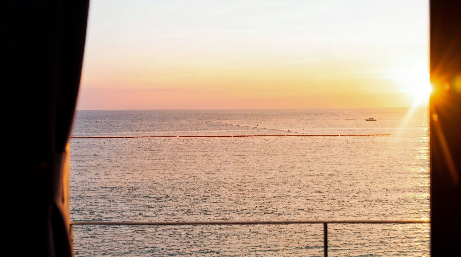 A view of the sea from a window with a railing, a boat in the distance, and the sun setting or rising.