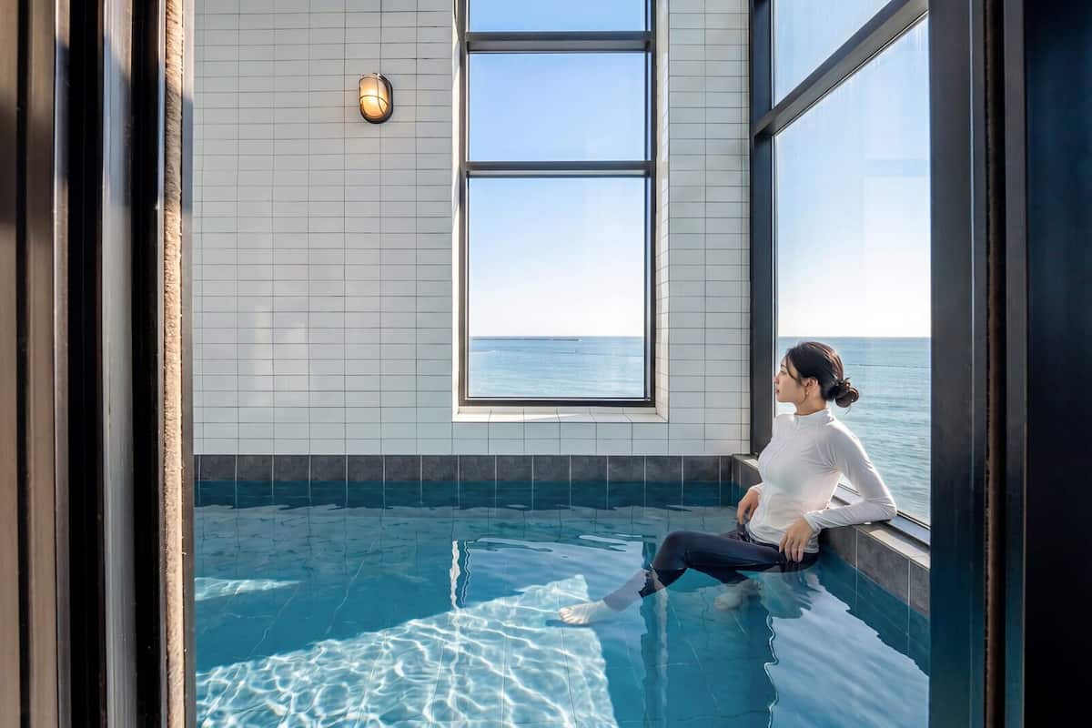 Una persona sentada junto a una ventana con vista a una piscina y al mar.