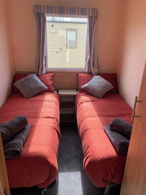 A compact room with two beds, a window with curtains, and a small table. - Cabin Sleeps 6 with Parking near Beach (New Romney)