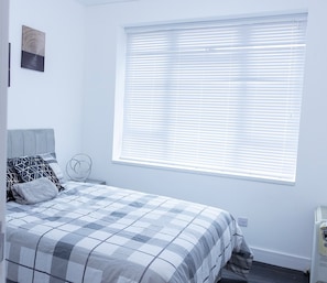 Ein Schlafzimmer mit Bett, einem Fenster mit Jalousien, einem Wandbild und einer Heizung.