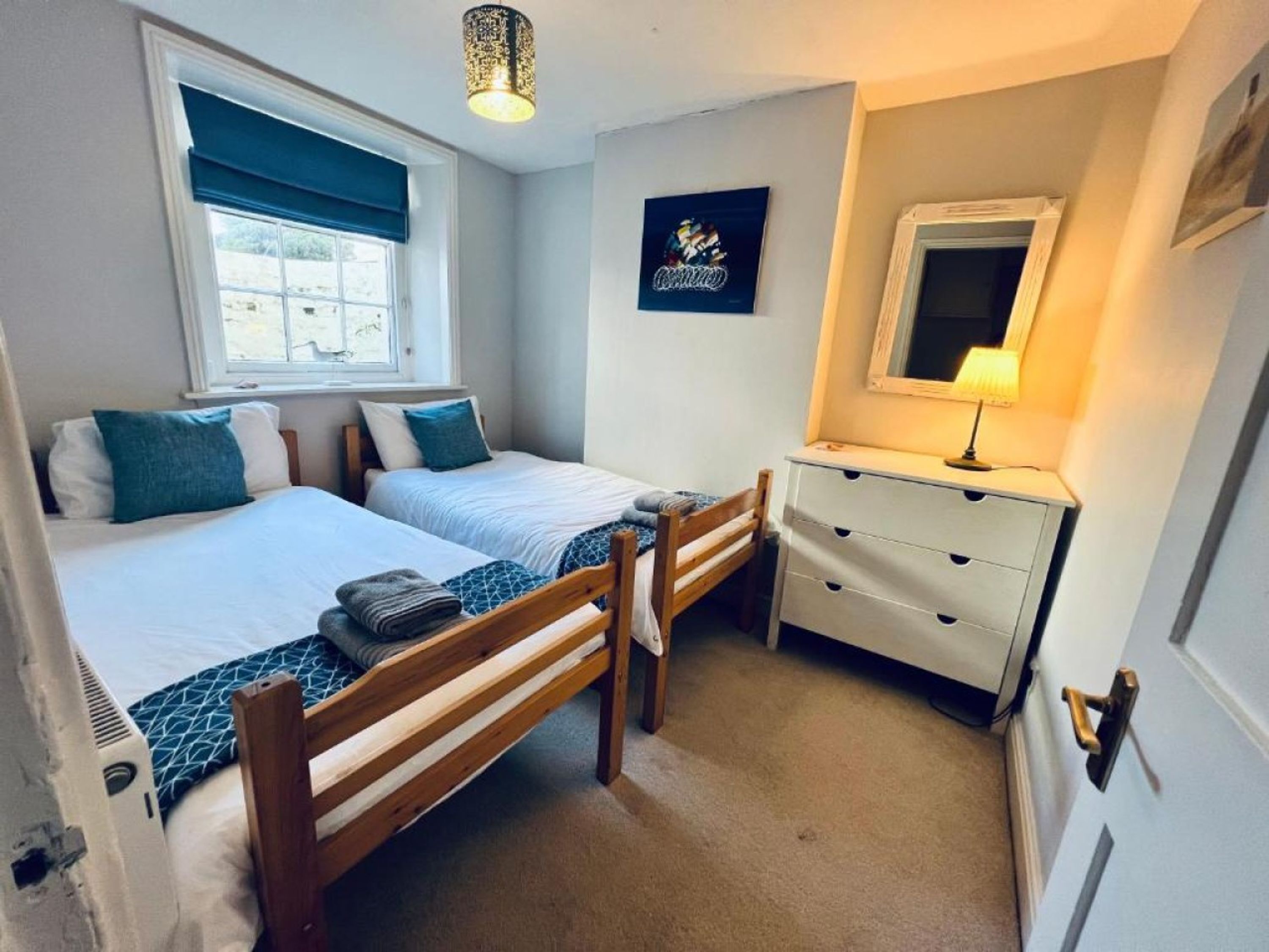 A bedroom with two beds, a dresser, a window, and a wall-mounted lamp.