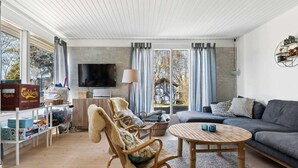 Ein Wohnzimmer mit Sofa, Sessel, Couchtisch, Fernseher und einem Fenster mit Vorhängen.