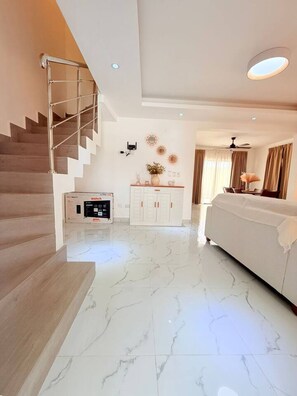 Una habitación de hotel moderna con piso de mármol, un sofá blanco, una escalera con barandillas de metal, un ventilador de techo y un televisor montado en la pared.