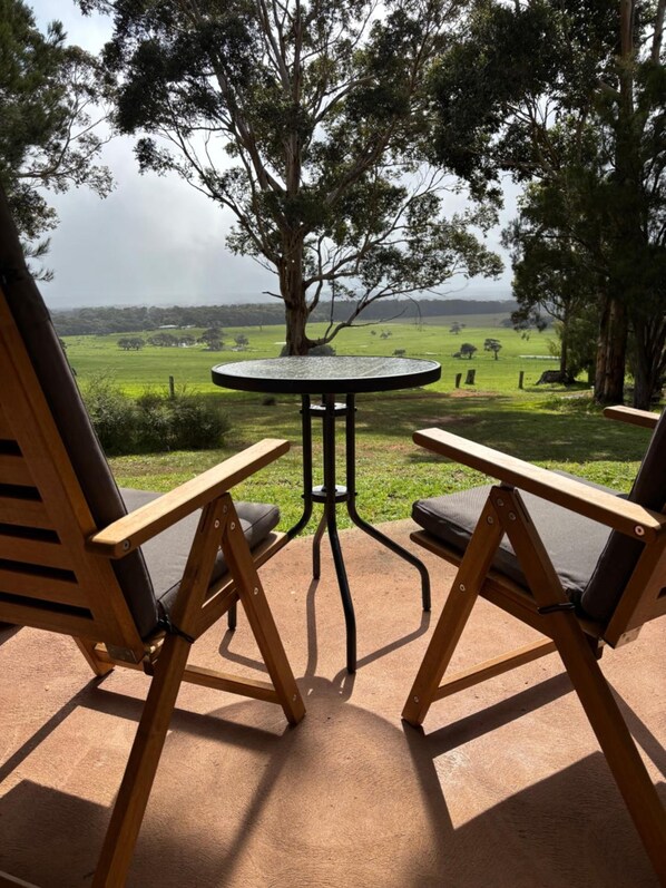 A patio with a round table and two chairs, overlooking a grassy field with trees.