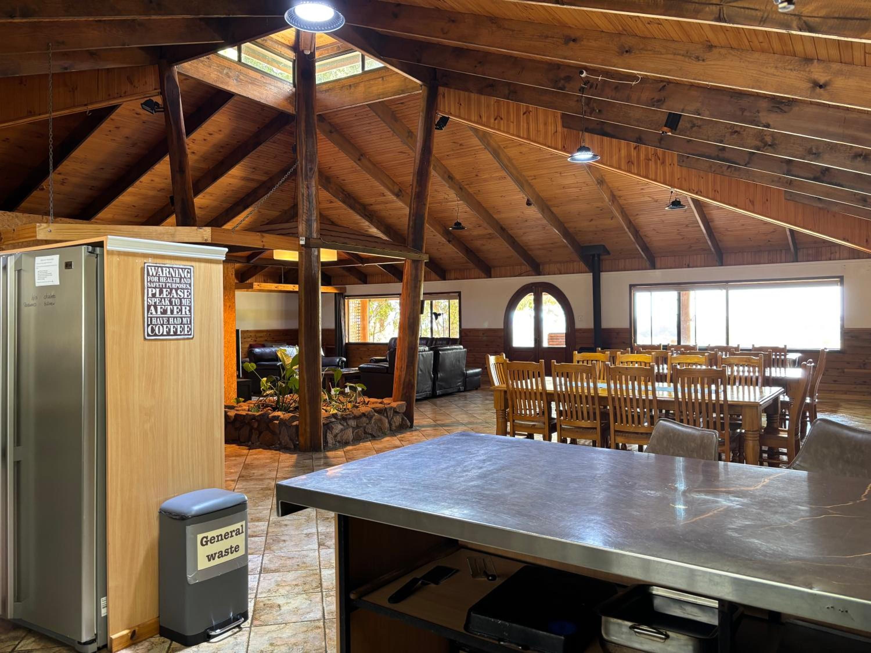 Un interior rústico con techo alto, vigas de madera, una zona de comedor con mesas y sillas, y una cocina con refrigerador y un basurero etiquetado como 'Residuos Generales'.