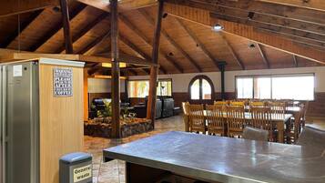 Un interior rústico con techo alto, vigas de madera, una zona de comedor con mesas y sillas, y una cocina con refrigerador y un basurero etiquetado como 'Residuos Generales'.