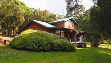Una casa de madera de dos pisos con techo verde, rodeada de abundante vegetación y árboles.