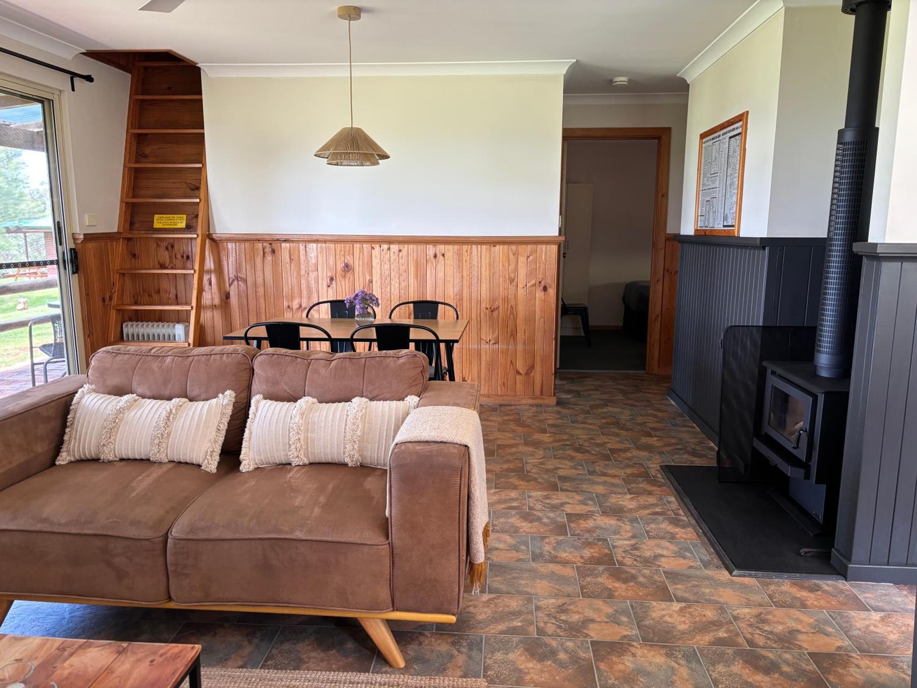 Ein gemütliches Wohnzimmer mit braunem Sofa, Holzvertäfelung, einem Esstisch mit Stühlen, einer Pendelleuchte und einem Kamin.