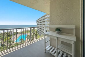 A balcony with a view of a pool and beach, a white shelf with a plant, and a building with balconies in the background. - Seaside Beach and Racquet 5617 | BeachFront + Pool (Orange Beach)