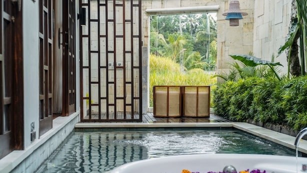 Une piscine intérieure paisible, avec vue sur une végétation luxuriante, une porte en bois et une baignoire blanche parsemée de pétales de fleurs.