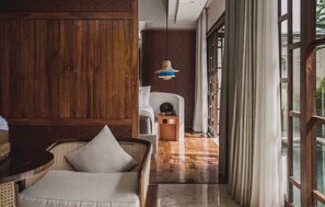 A modern interior with a wooden panel wall, a wicker sofa with a cushion, a hanging light fixture, and a view through a glass door. - asvara villa (Ubud)