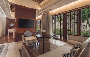 A modern living room with a flat-screen TV, comfortable seating, a round table, and large windows with a view of greenery.