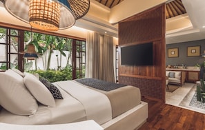 A bedroom with a bed, a TV mounted on a wooden panel, a desk with a chair, and a view of greenery outside.