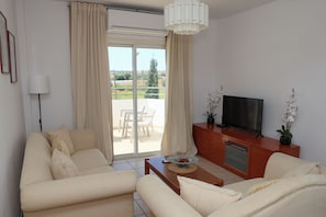 A living room with a sofa, a coffee table, a television on a wooden cabinet, and a view through a sliding glass door.