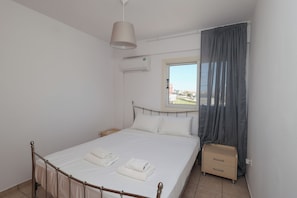 A neatly made bed with white linens and towels, a bedside table with a lamp, a window with curtains, and an air conditioning unit on the wall.