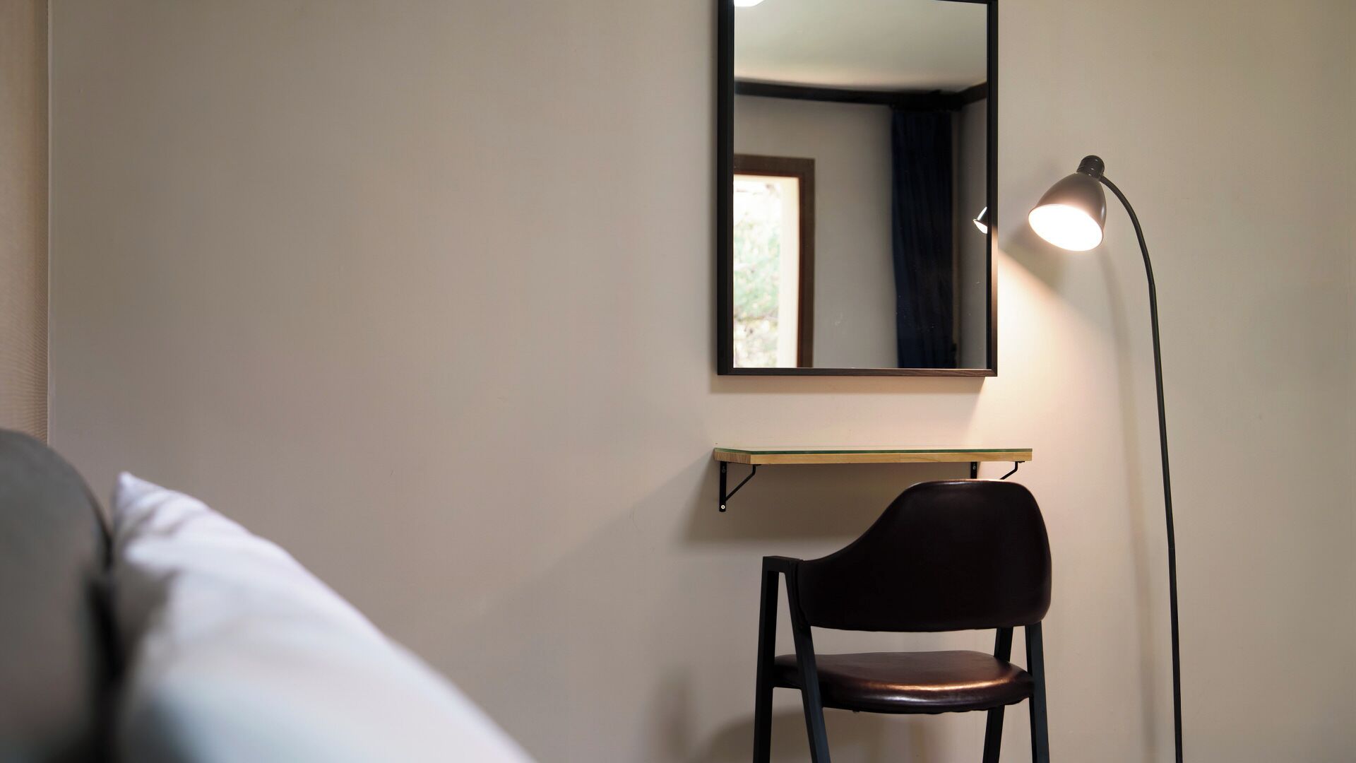 A room with a bed, a chair, a desk with a mirror, and a floor lamp.