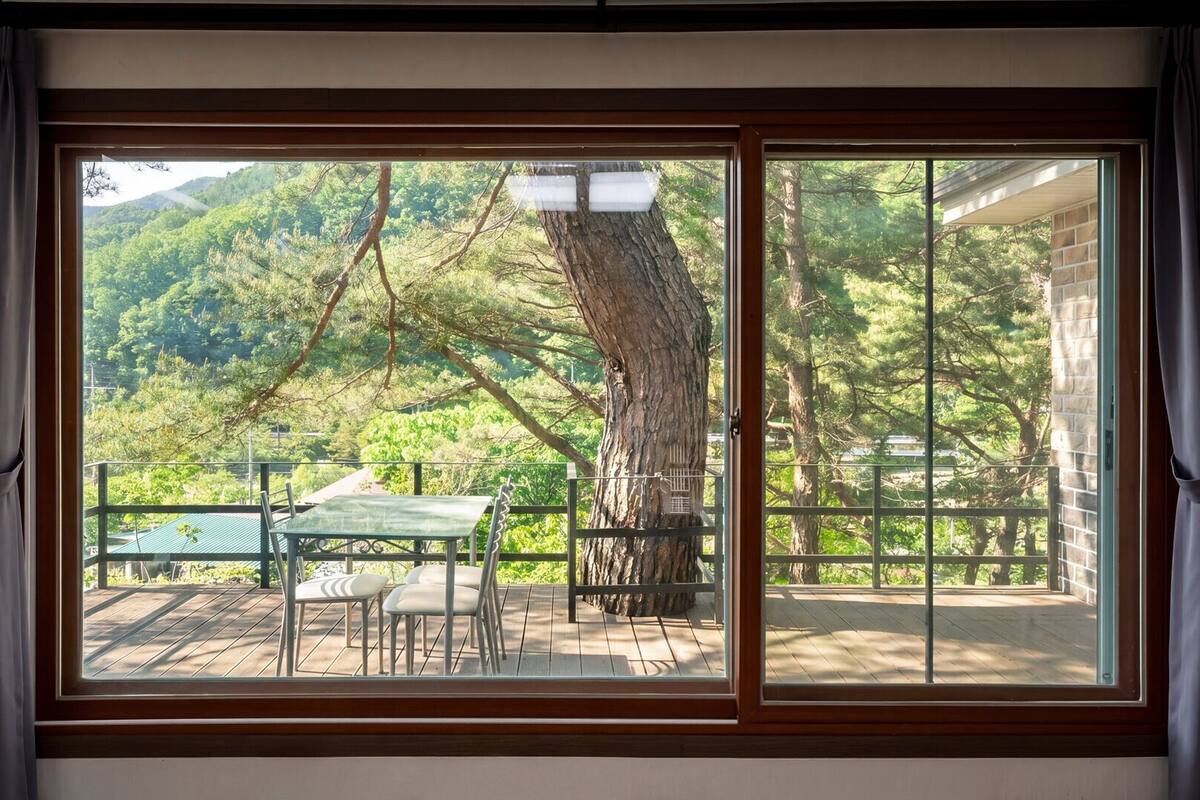 Una habitación con un amplio ventanal corredera que da a una terraza exterior con mesa y sillas, rodeada por una valla de madera y un gran árbol.