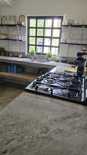 A kitchen with a gas stove, a window with a view of greenery, and various kitchen items on shelves. - "1200 m² Bacalar: Laguna a Pasos, Pet Friendly" (Xul-Ha)