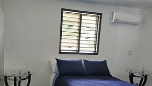 A bedroom with a bed, a window with blinds, a wall-mounted air conditioner, and a small table.