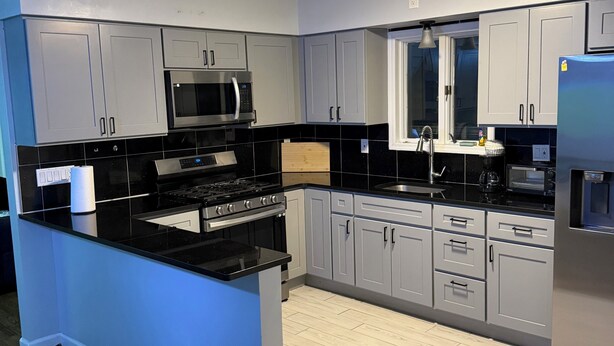 A modern kitchen with grey cabinets, a black countertop, a stainless steel refrigerator, and a built-in microwave. - Modern 3BR Jersey City Retreat Near NYC & FIFA (Jersey City)