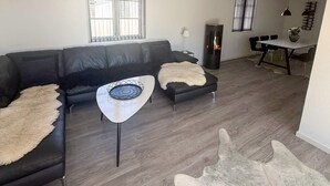 A modern living room with a black leather sofa, a white coffee table, and a fur throw. There is a fireplace, a dining table with chairs, and a large window with blinds.