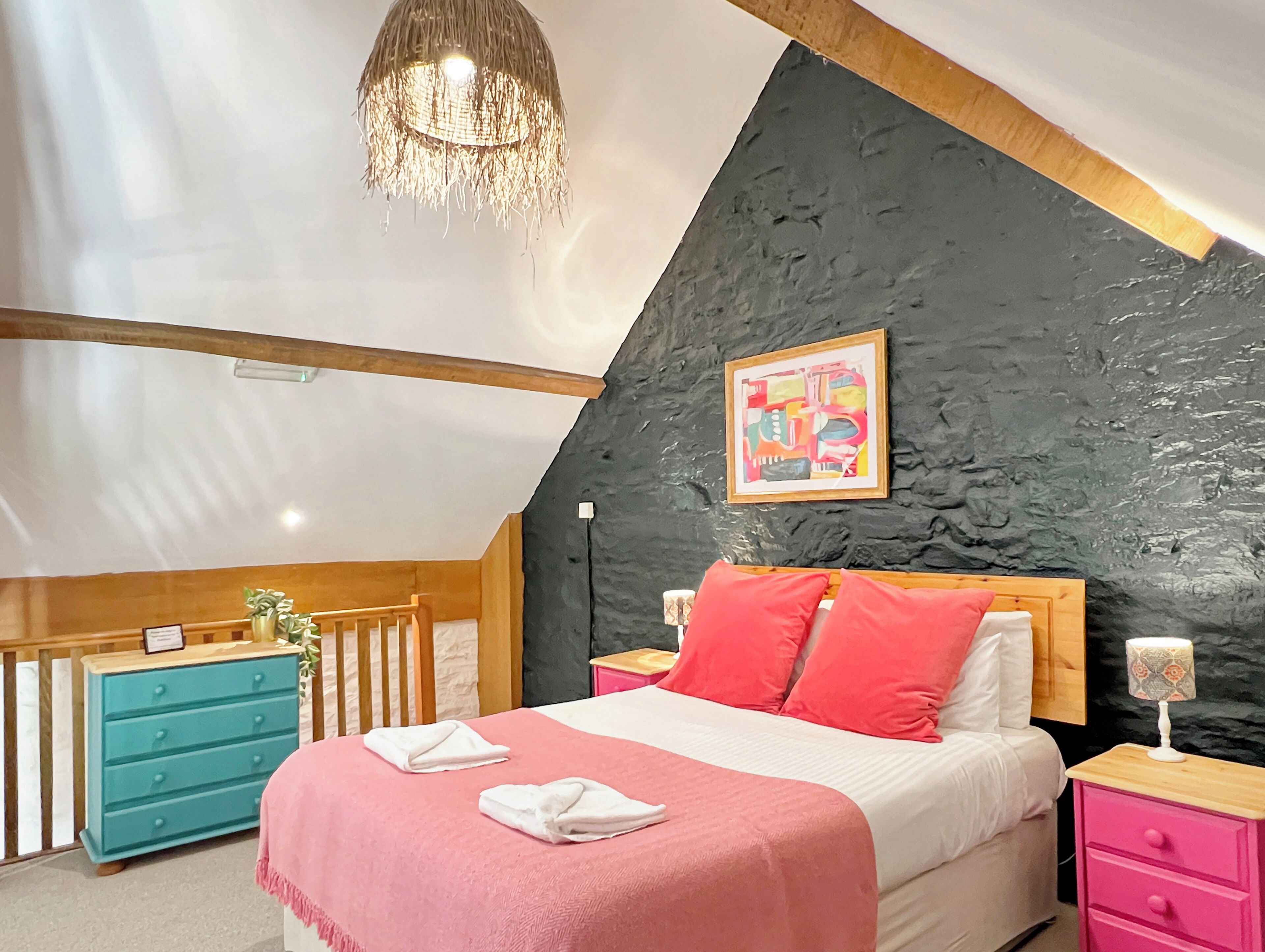 A bedroom with a sloped ceiling, a bed with pink and white bedding, a blue dresser, a wooden nightstand, and a wall-mounted picture.