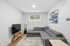Ein modernes Wohnzimmer mit einem Ecksofa, einem Flachbildfernseher, einer Holz-TV-Konsole, einem kleinen Tisch, einem gemusterten Teppich und einem Bild an der Wand.