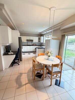 Cozinha moderna com armários brancos, sala de jantar com mesa redonda e cadeiras, e vista para o exterior através de uma porta de correr de vidro.