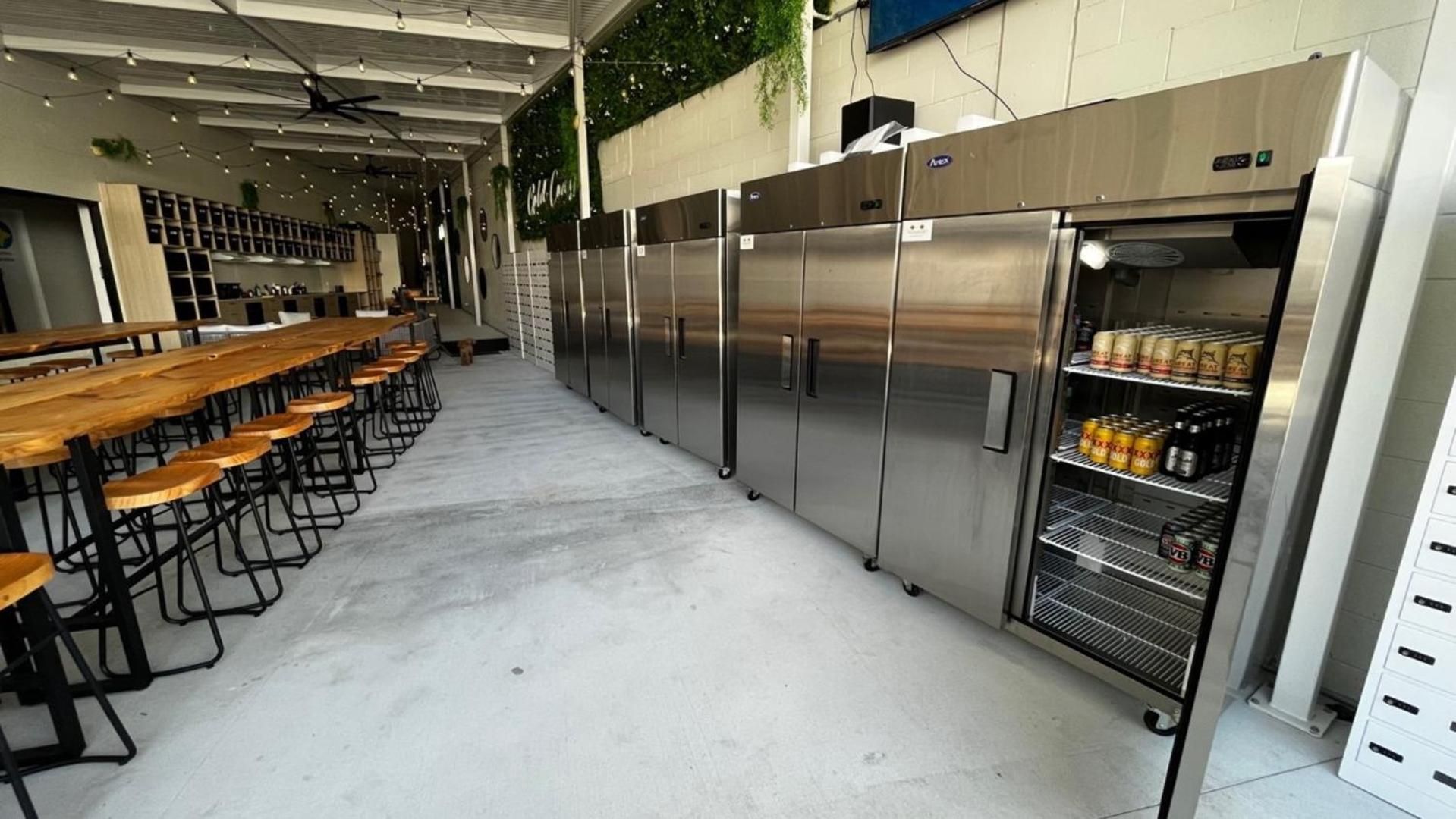 A commercial kitchen with stainless steel appliances, a long wooden table with stools, and a refrigerated display case.