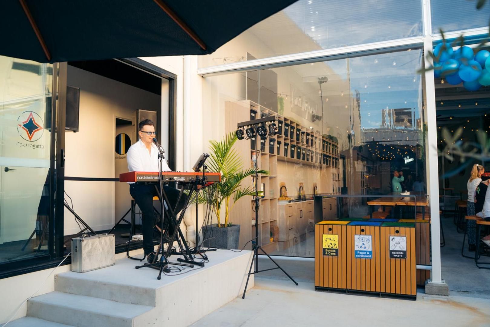 A person playing a keyboard outside a building with a glass wall, a trash bin, and a sign for 'Bottles & Cans'.