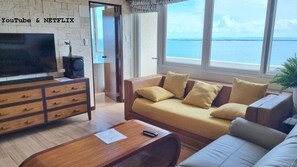 A living room with a sofa, a wooden coffee table, a TV, and a speaker. There is a view of the sea through large windows. - Huge Lux. 2 BR Condo (100 SQM)  2 pools - Beach, Sea-views, Gym, Tours available (Lapu-Lapu City)
